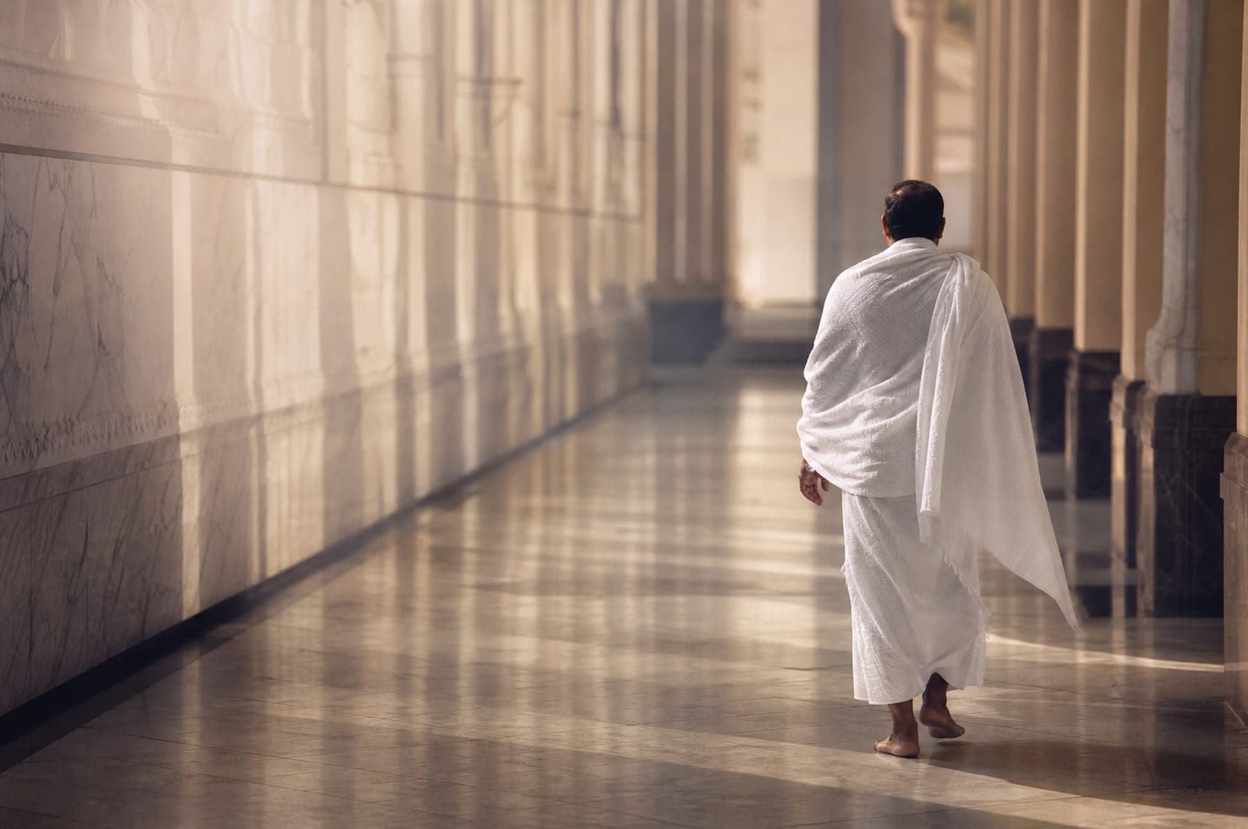 A pilgrim in ihram walking through a shaded marble colonnade in early morning light