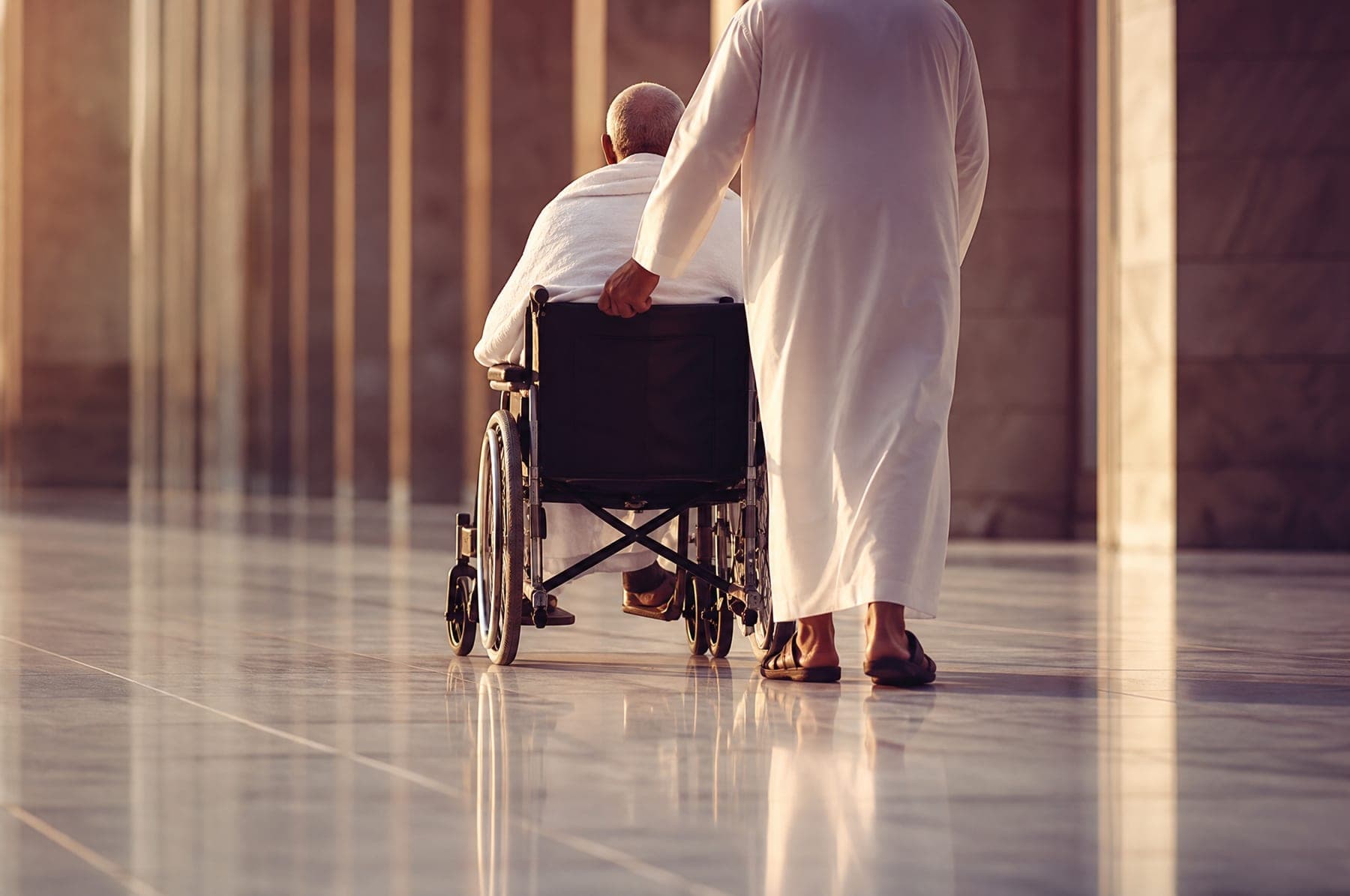 An elderly pilgrim in a wheelchair being pushed along smooth marble by an attendant